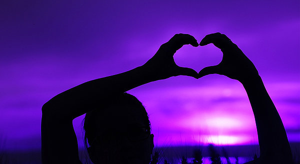 Eine Frau zeigt im Sonnenuntergang ein Herz mit einer Hand